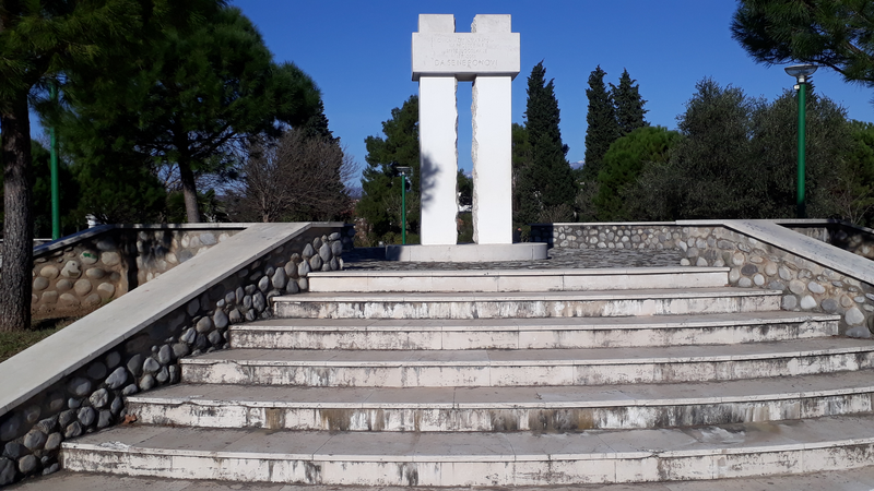 Platform with stairs up to white monument consisting of two pillars and cross-piece with the inscription: "Civilnim žrtvama ratova na prostorima bivše Jugoslavije 1991--2001 DA SE NE PONOVI"
