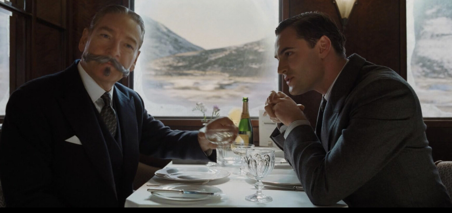 Kenneth Branagh as Hercule Poirot in Murder on the Orient Express (2017), seated in the dining car, looking into the camera. (Flipped horizontally from original.)