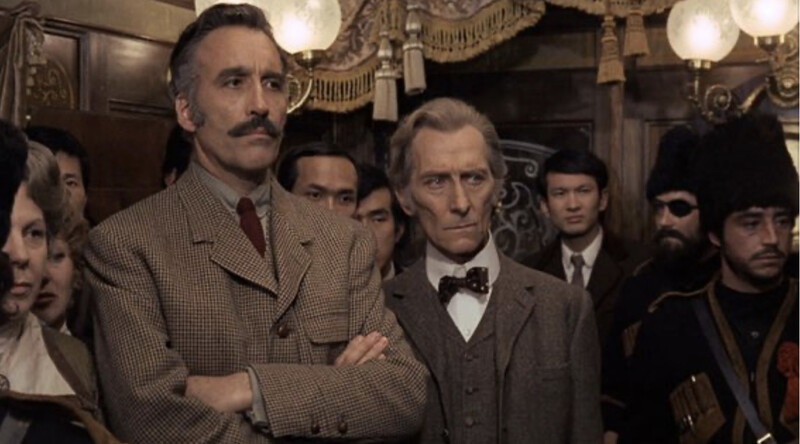 Peter Cushing and Christopher Lee in Horror Express (1972), standing in an ornate but crowded train car with various Eastern-looking characters, looking sombre and perhaps a bit impatient