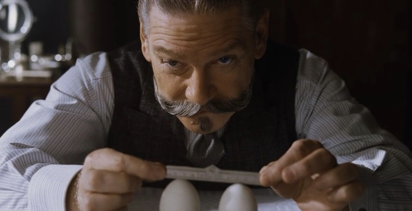 Branagh as Poirot in Murder on the Orient Express (2017), bent over the table, measuring two eggs at the tip with a ruler
