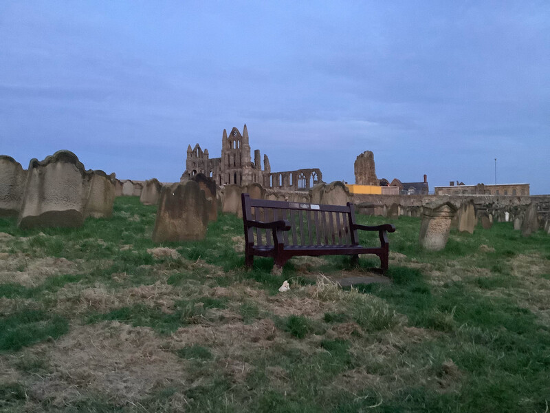 Bench in St Mary's churchyard, Whitby, surrounded by old tombstones in the dwindling light. Above, against the deep dusk of the sky, Whitby Abbey is seen.