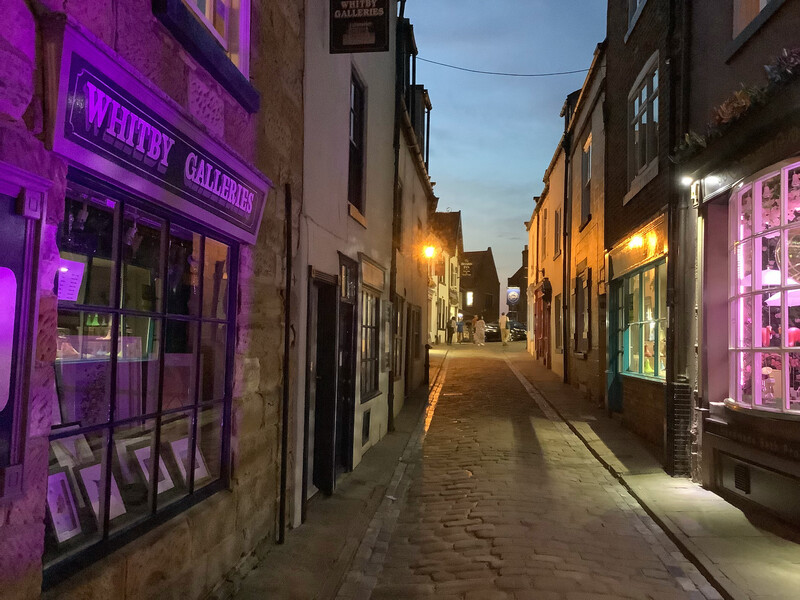 Church Street, Whitby, at night. Narrow, with cobblestones. The display window of Whitby Galleries close up on the left.