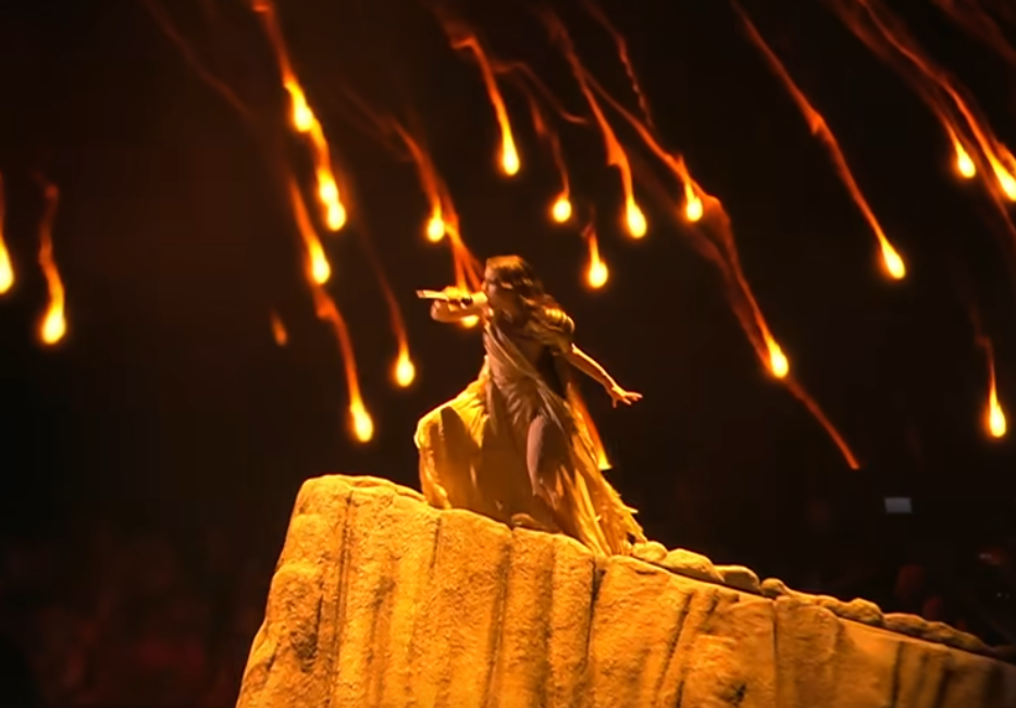 Slika z odra. Pevka na klancu, z naramnim oklepom nad dolgo obleko iz muslina, pod nebom s padajočimi plameni belega fosforja.